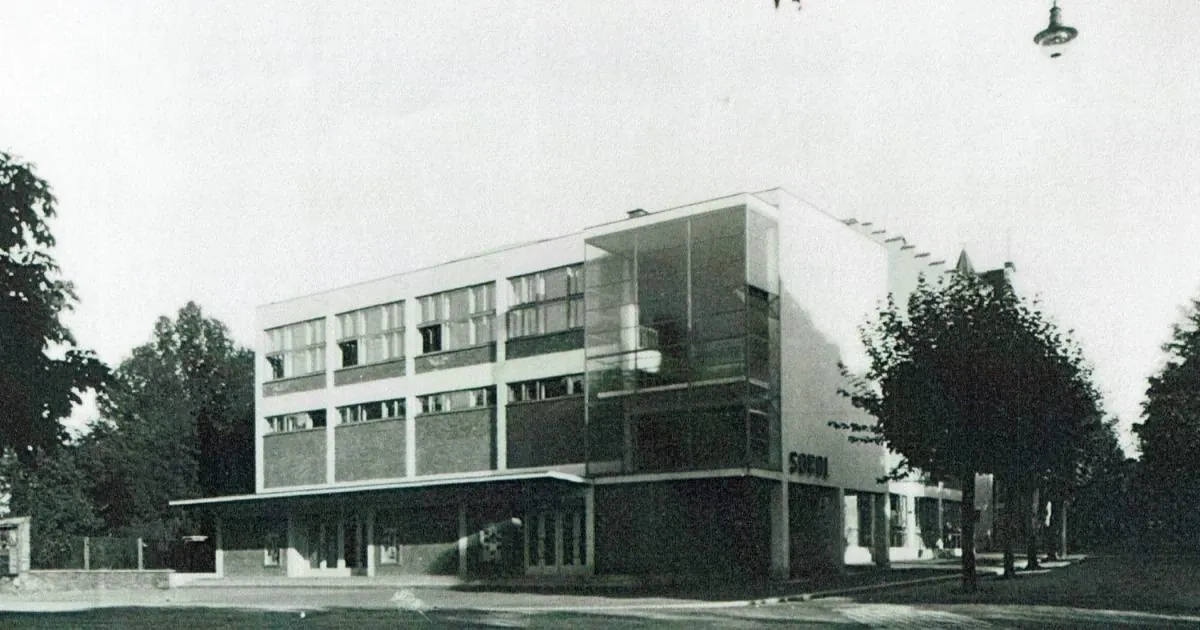 Dobová černobílá fotografie funkcionalistické budovy sokolovny s nápisem SOKOL, před ní ulice a stromy.
